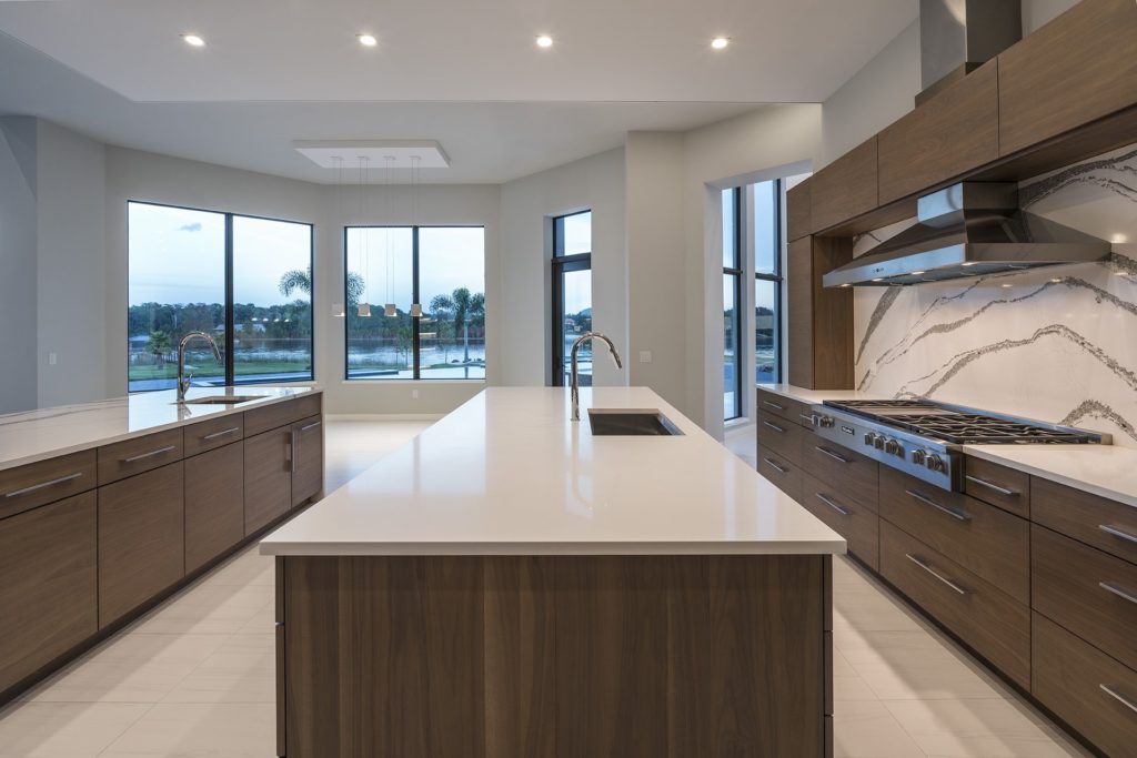 The kitchen backsplash crafted of white quartz with gray veining echoes the wraparound countertop of the island facing the great room in this Florida Modern home by Orlando Custom Homebuilder Jorge Ulibarri 