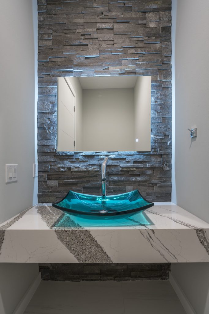 Powder Bath with blue glass vessel sink that glows from LED lighting and sits atop a white and gray quartz floating vanity framed by a floor-to-ceiling ledgestone wall