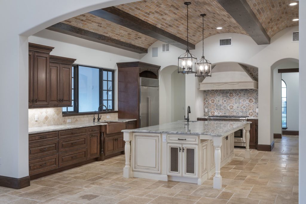 The granite island counter and granite perimeter counters with gray and white veining contrast with walnut wood stained cabinetry and travertine floors and backsplashes to give this kitchen its Spanish Revival style in this custom home by Orlando custom homebuilder Jorge Ulibarri.