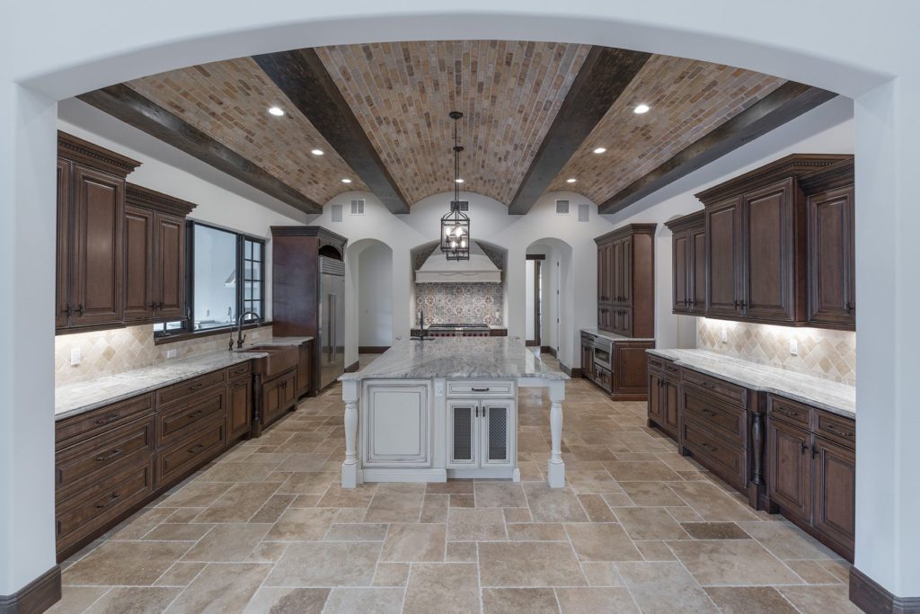 The kitchen opens to the great room and features a brick and beam barrel ceiling with a contrasting ivory colored island and handpainted tile backsplash in this Spanish Revival Custom Home by Orlando Custom Homebuilder Jorge Ulibarri.
