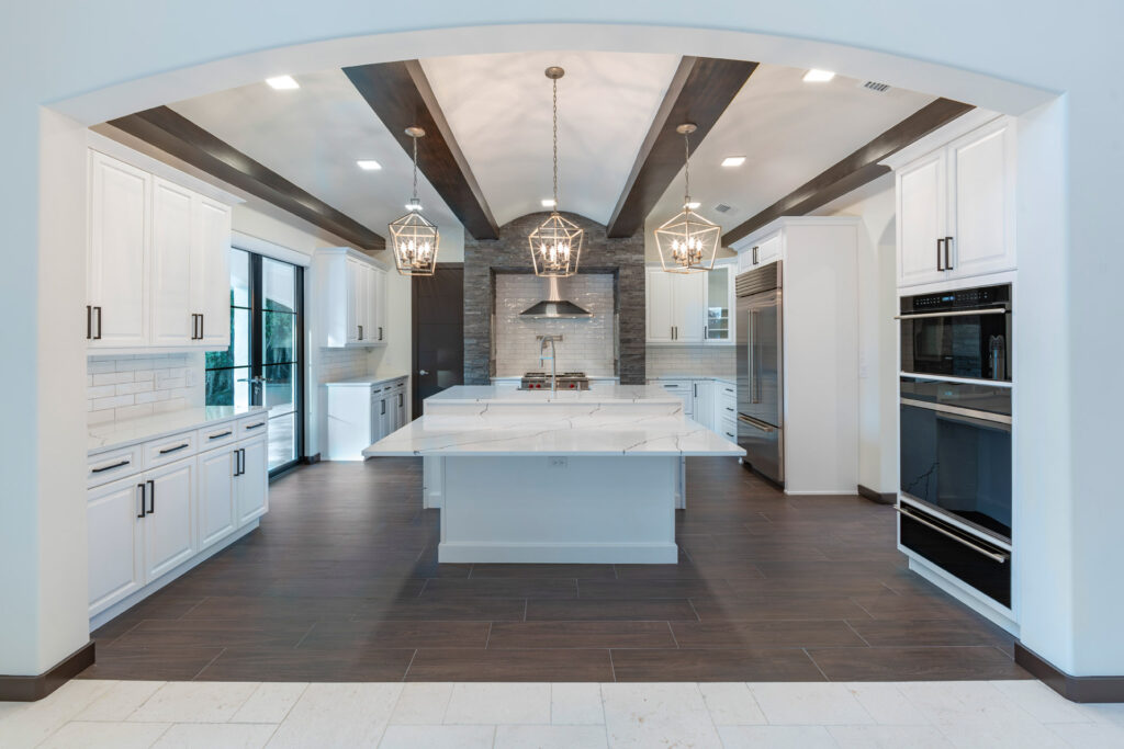 The open kitchen is sleek with white cabinets and quartz counter tops grounded with wood elements including the ceiling beams and the porcelain wood plank floors.