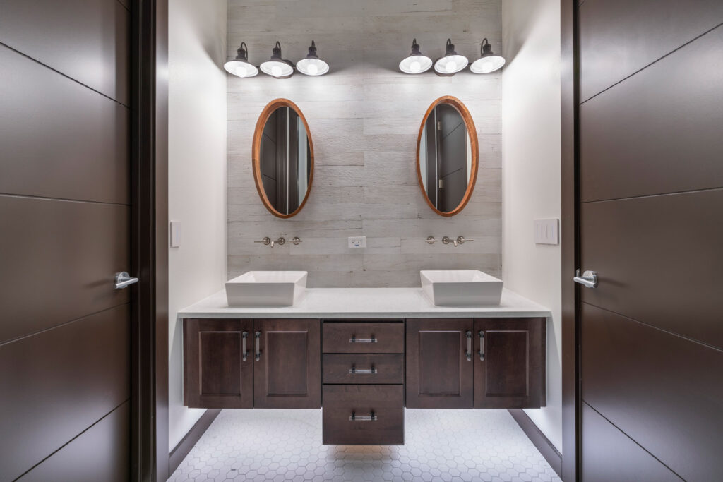 This bathroom conveys a more rustic vibe with its industrial style lighting fixtures and distressed porcelain gray wood plank accent wall behind the vanities with porcelain octagon mosaic tile floors.